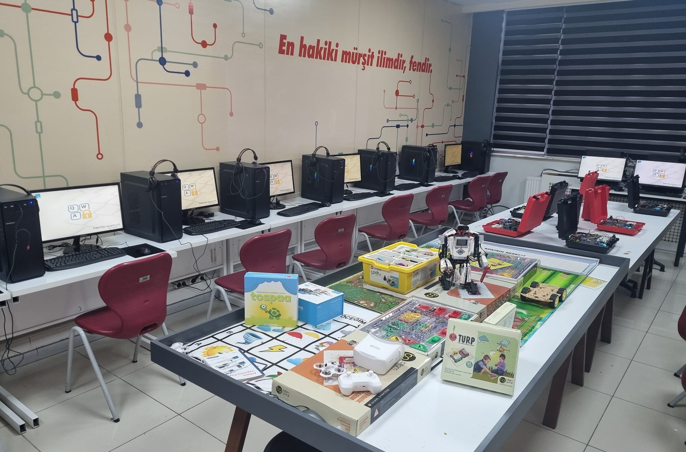 Bilişim sınıfında robotik ve kodlama masası: insansı robot seti, kodlama oyun pistleri, devre setleri ve küçük bir drone; arkada kulaklıklı bilgisayarlar, duvarda “En hakiki mürşit ilimdir, fendir.” yazısı.