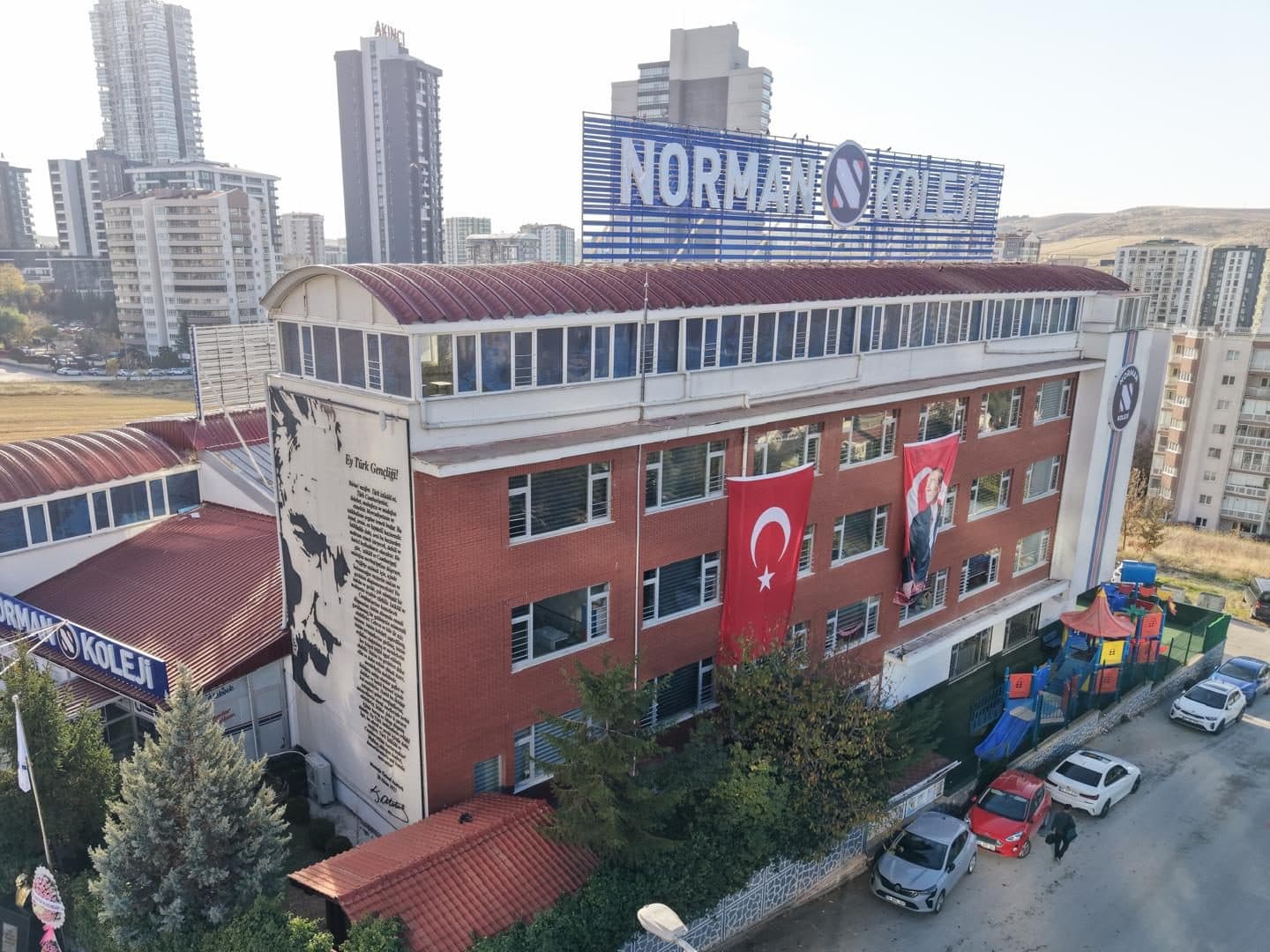 Norman Koleji binası önden, yakın havadan: kırmızı tuğla cephede Türk bayrağı ve Atatürk posteri, çatıda NORMAN KOLEJİ tabelası, solda Atatürk'ün Gençliğe Hitabesi duvar resmi, sağda oyun parkı.