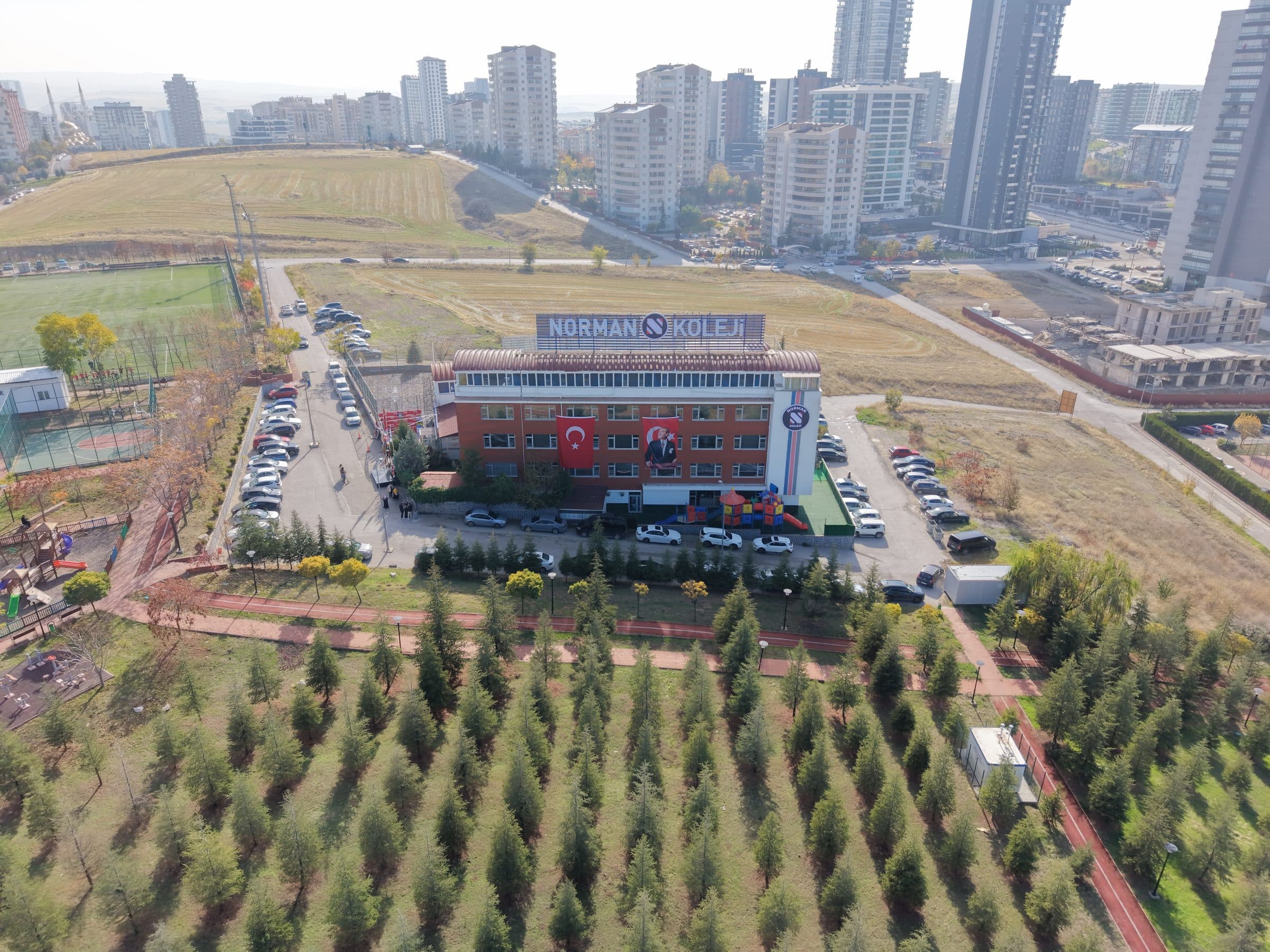 Norman Koleji binasının havadan görünümü: çatısında NORMAN KOLEJİ tabelası olan kırmızı tuğla cepheli bina, önünde ağaçlık alan ve otopark, arkada Yaşamkent'in yüksek konutları.