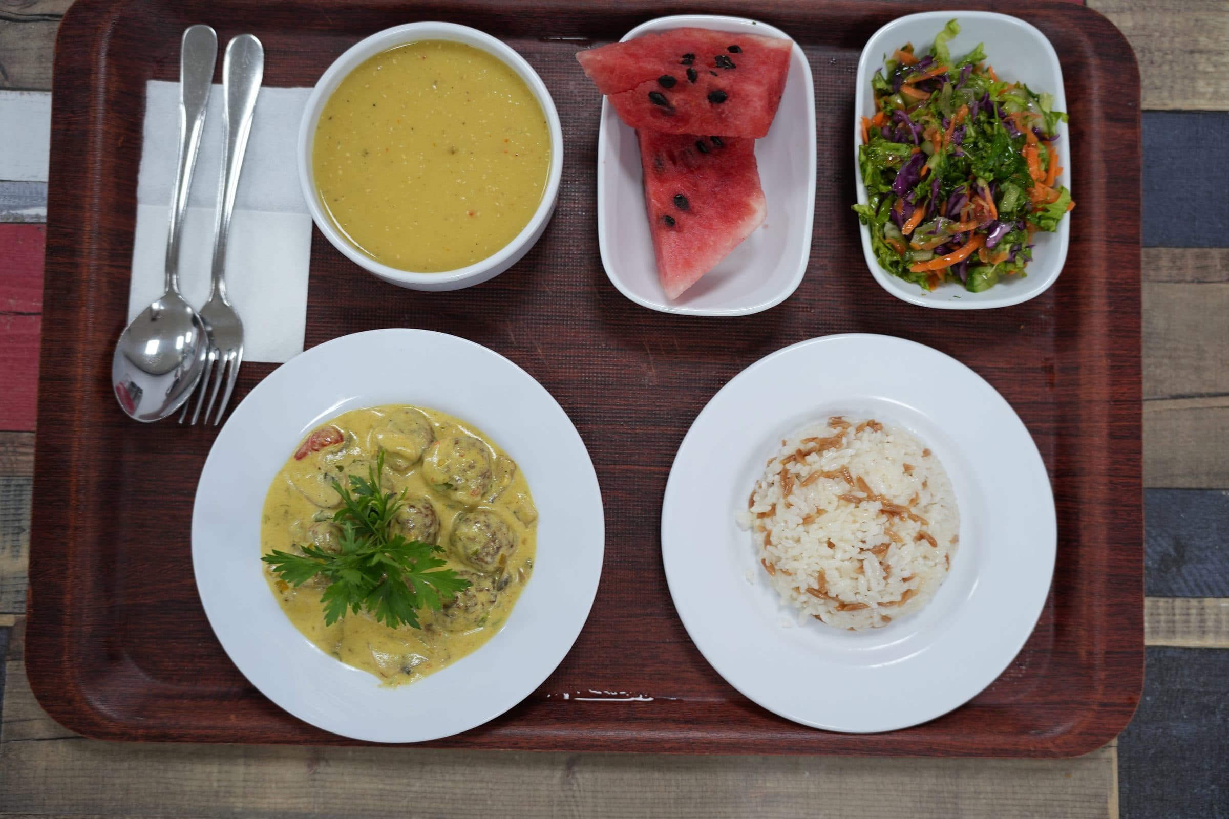 Okul yemekhanesinden bir öğle yemeği tepsisi: çorba, ana yemek, pilav, salata ve karpuz.