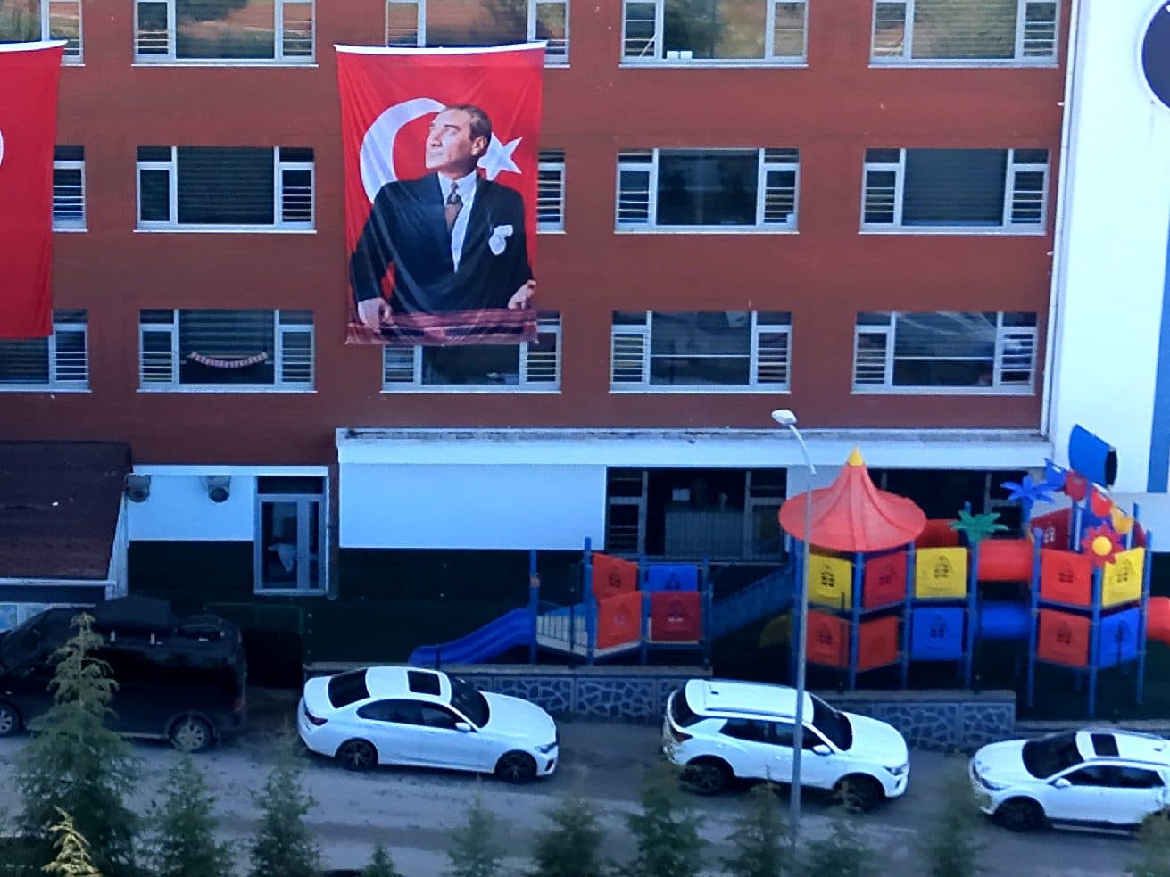 Okul binasının önünde suni çimli alanda renkli çocuk oyun parkı: kaydıraklar, kuleler ve tüneller; tuğla cephede Türk bayrağı ve Atatürk posteri.