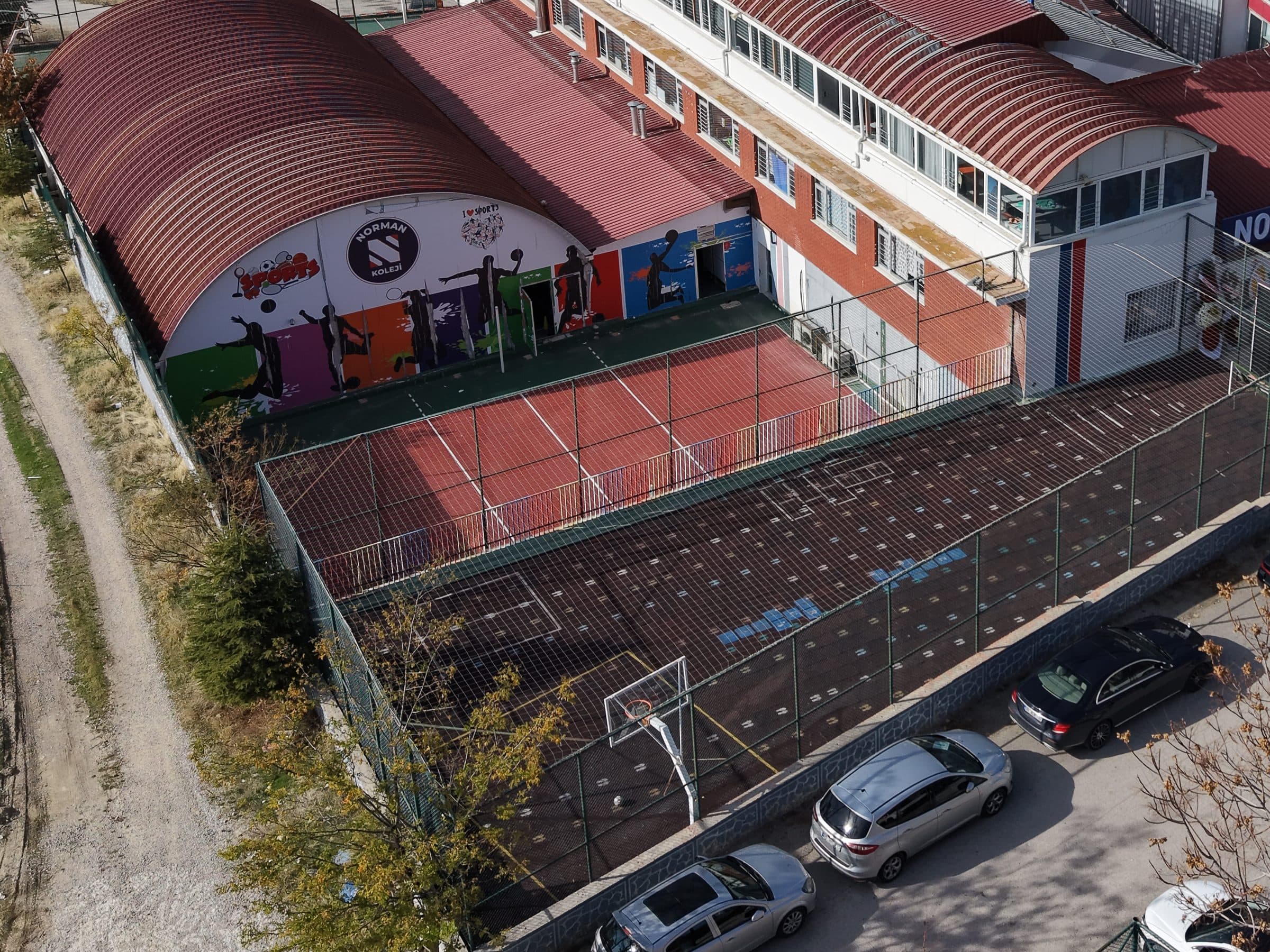 Havadan görünüm: kemer çatılı spor salonunun spor temalı duvar resmi olan cephesi önünde, tel çitle çevrili iki açık saha; biri kırmızı zeminli ve kaleli, öteki basketbol potalı.