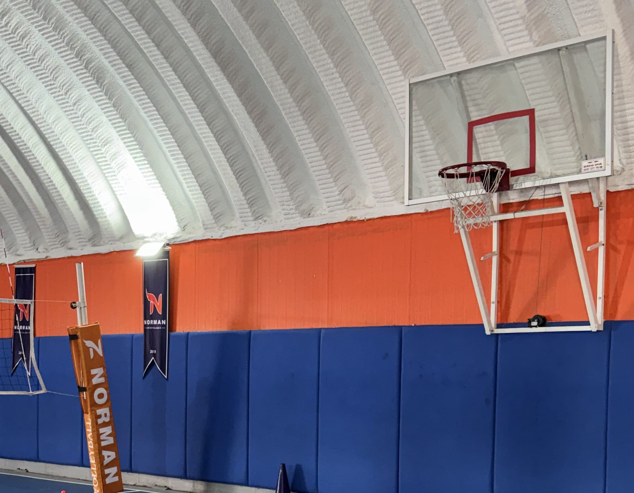 Okulun kapalı spor salonu: beyaz kemerli tavan, turuncu ve lacivert duvar, cam panolu basketbol potası, voleybol filesi ve duvarda Norman Spor Kulübü flamaları.