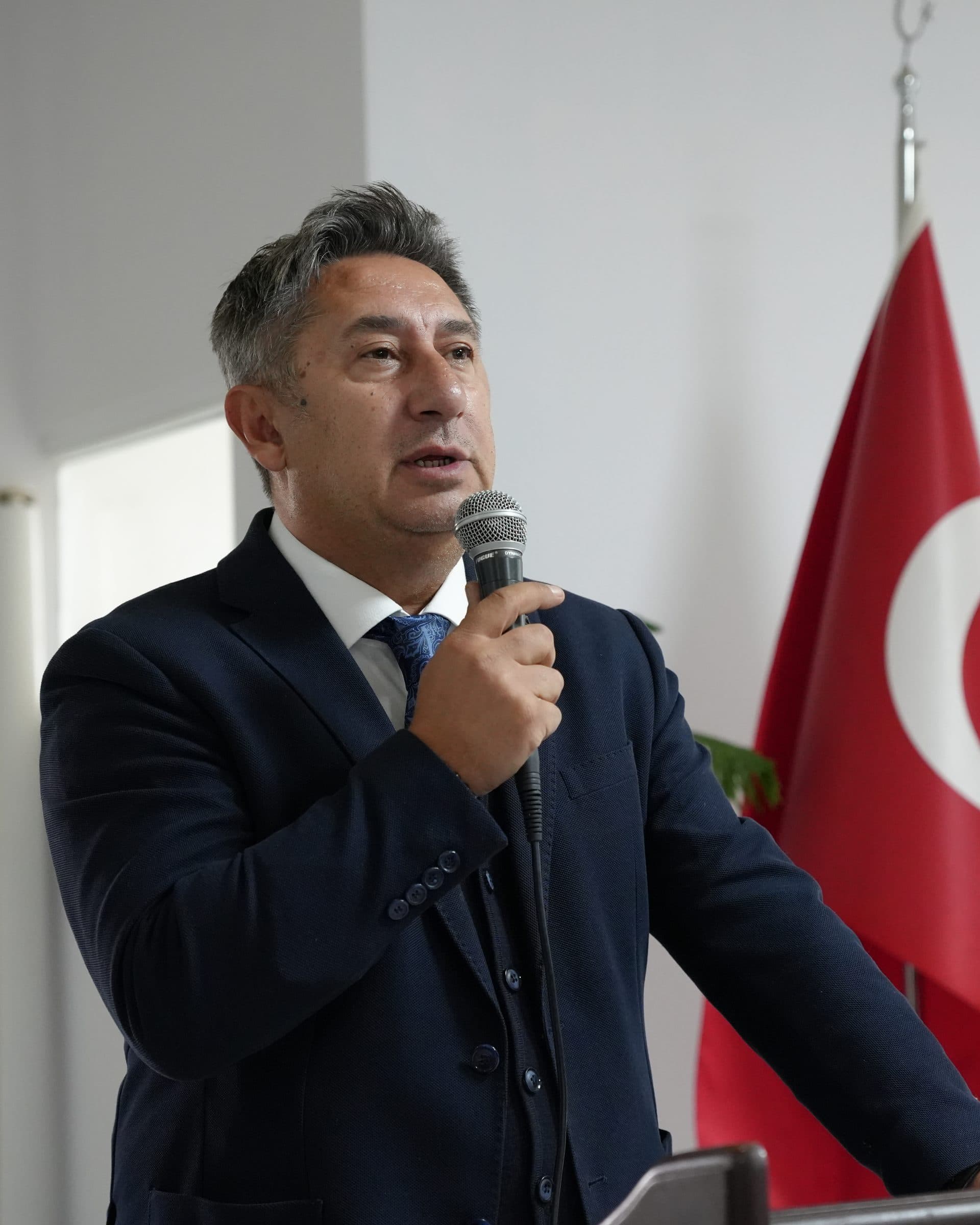 Öztuna Norman speaking at the lectern at the school's opening on 14 September 2026; a Turkish flag behind him.