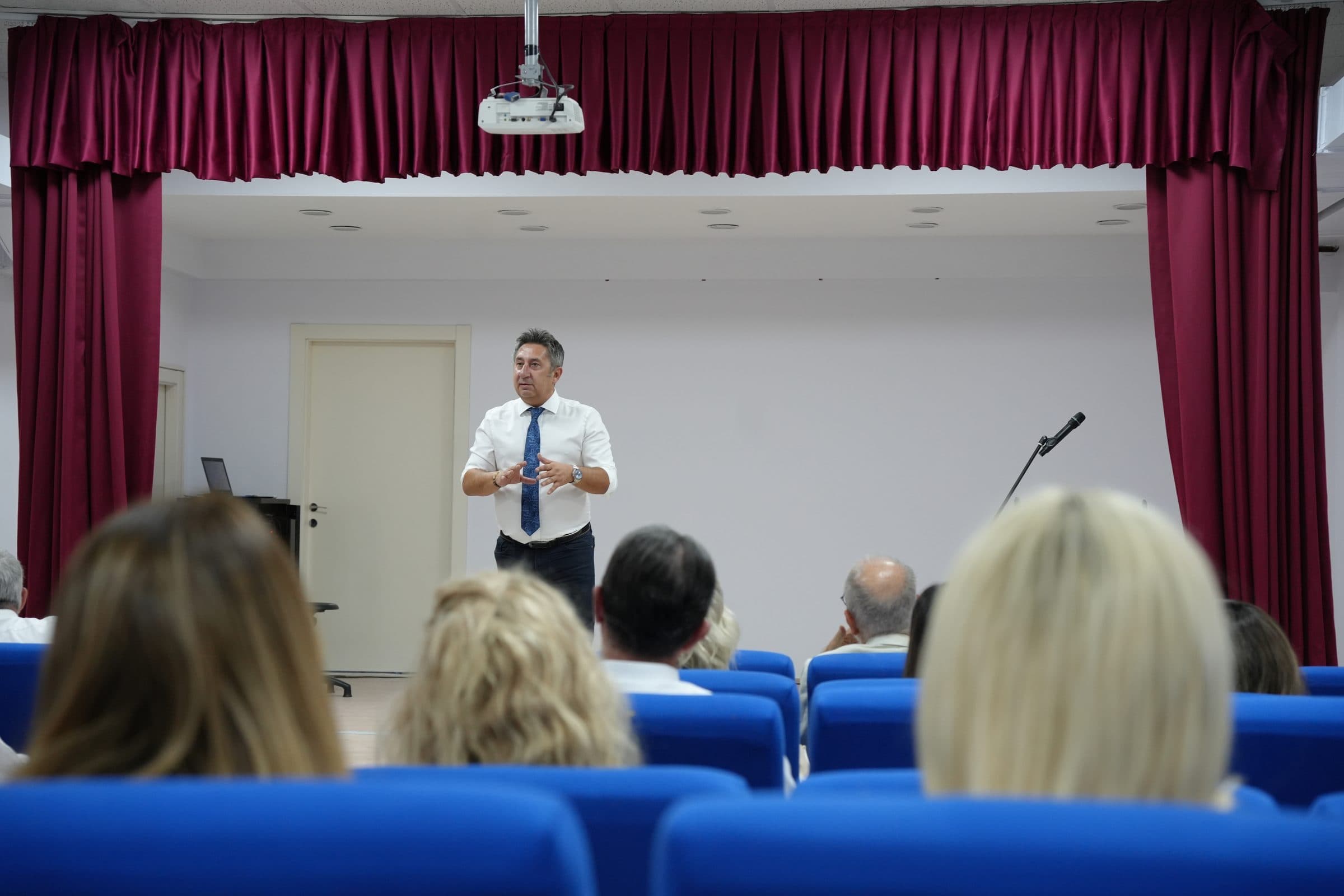Konferans salonunda mavi koltuklarda oturan öğretmenler, sahnenin önünde konuşan Öztuna Norman'ı dinliyor.